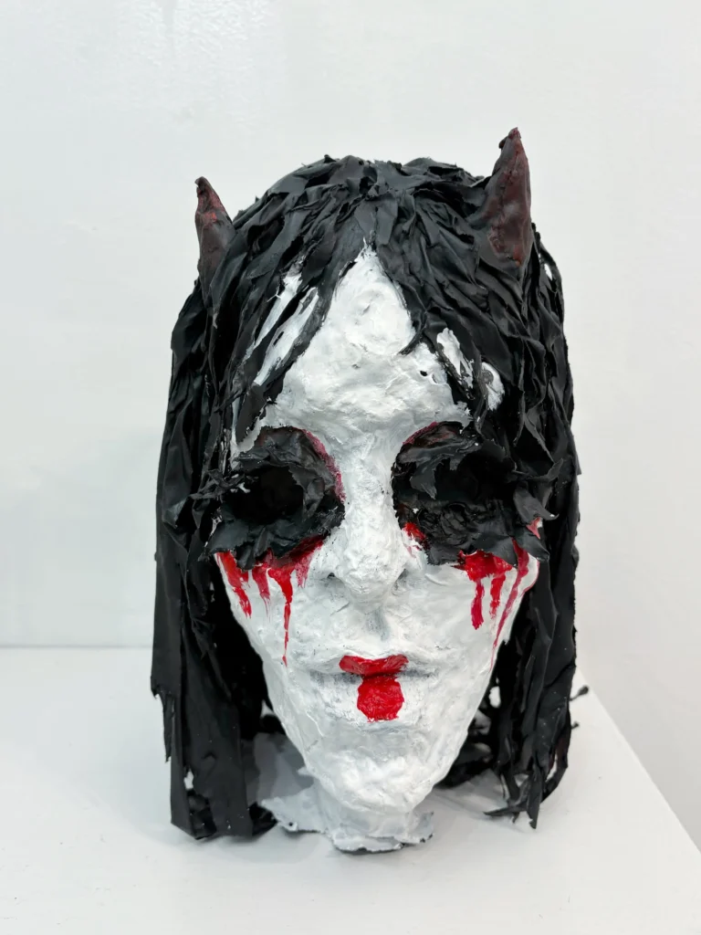 A papier-mâché Akuma mask: white face, black paper hair, small horns